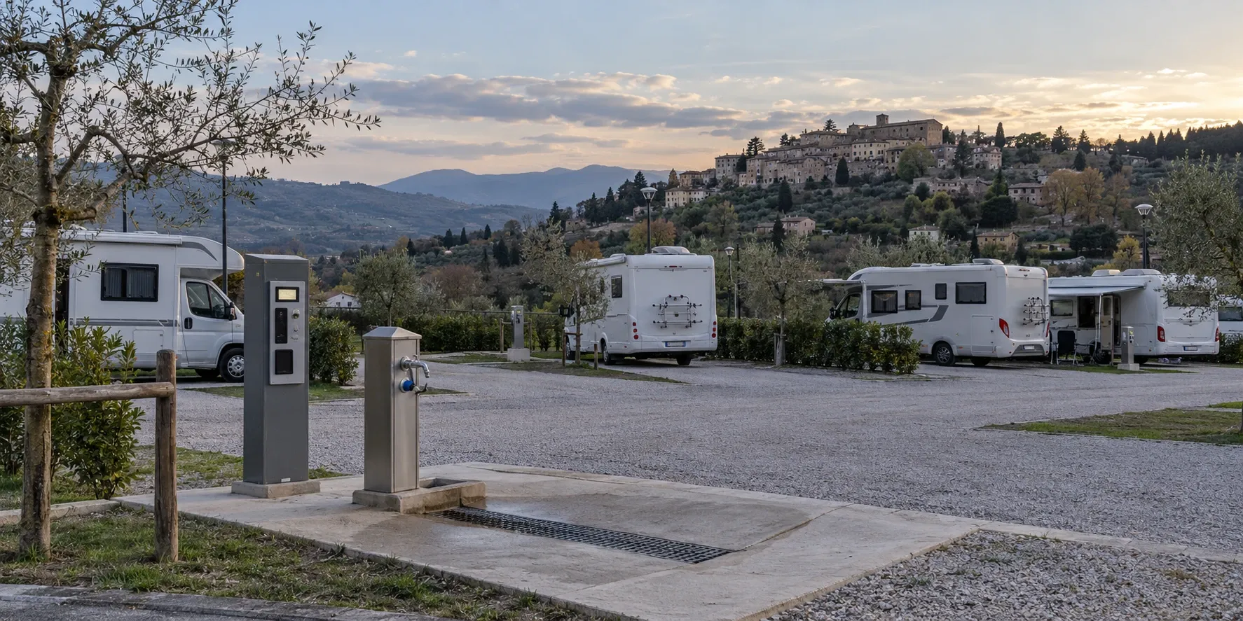 Area di sosta camper con colonnina di pagamento e punto di carico e scarico, borgo collinare sullo sfondo